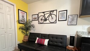 A living room with a black sofa, a potted plant, framed pictures on the wall, and a kitchen area with a countertop and appliances. - LA CASITA ON EAST 7TH STREET (Austin)