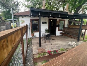 Un piccolo edificio rustico con una terrazza in legno, un'area salotto coperta e un giardino recintato.