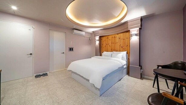 Ein modernes Schlafzimmer mit einem großen Bett, einem Holzkopfende, einer runden Deckenleuchte, einem kleinen Tisch und einem Stuhl.