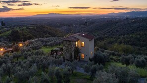 Uma casa rodeada por oliveiras numa encosta, com um pôr do sol ao fundo.