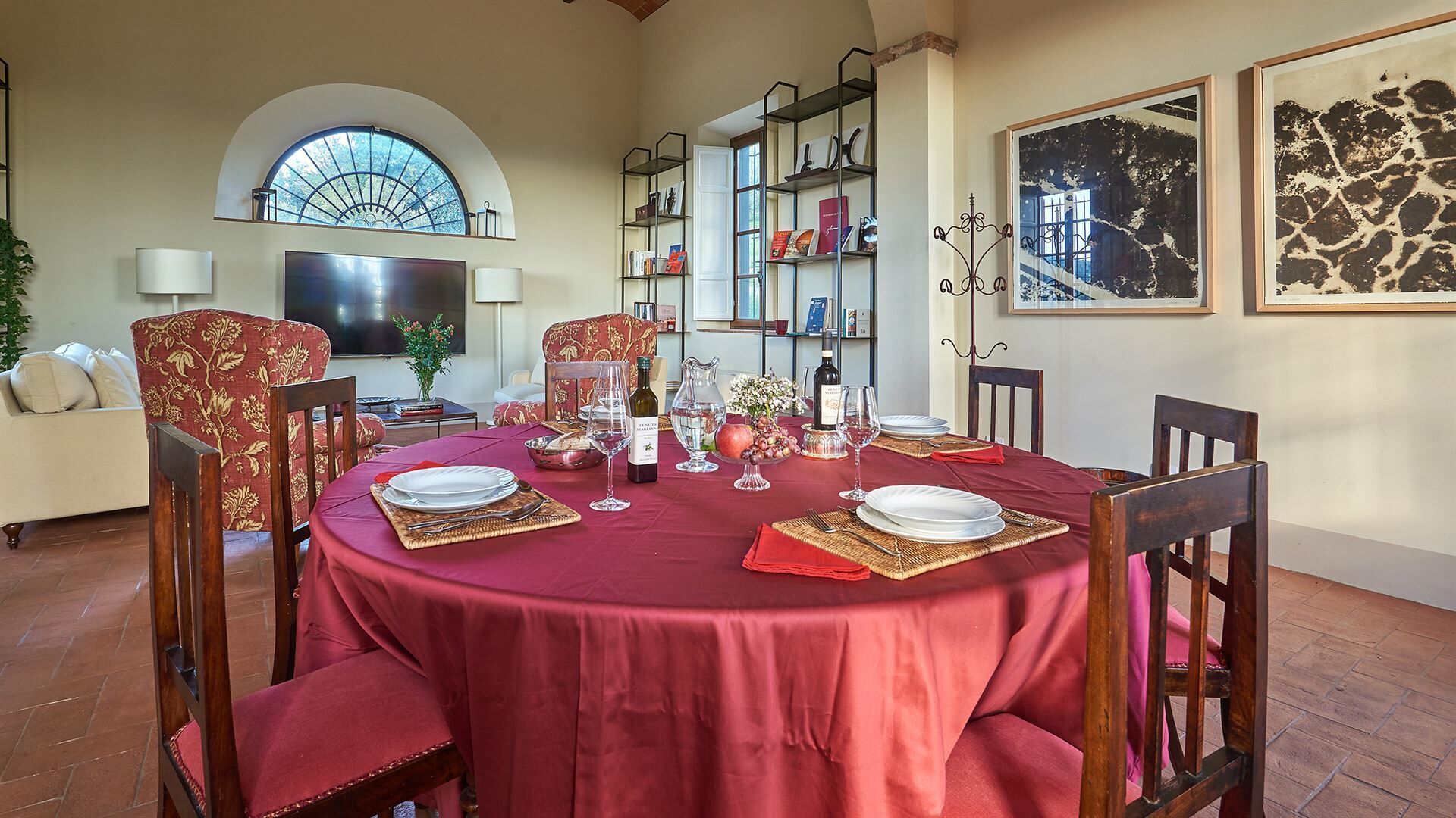 A dining area with a round table set for six, surrounded by chairs. There are bookshelves against the wall, a television, and a large window with a view of trees.
