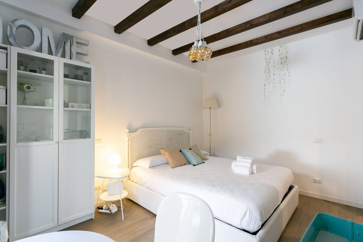 A bedroom with a bed, a white wardrobe, a small table with a lamp, and a hanging plant.