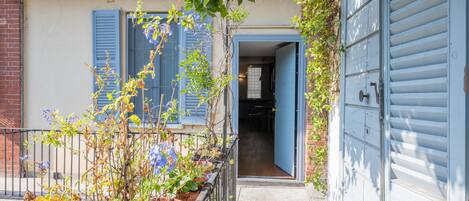 A narrow pathway leading to a building with a blue door and shutters, surrounded by potted plants and a metal railing.