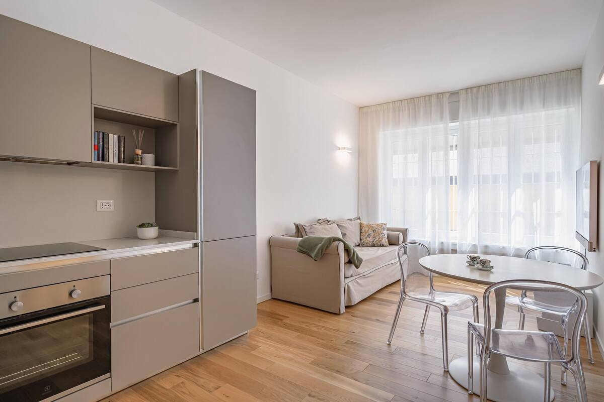 A modern kitchen with built-in appliances, a dining area with a round table and chairs, and a sofa with a view of the outside through large windows.