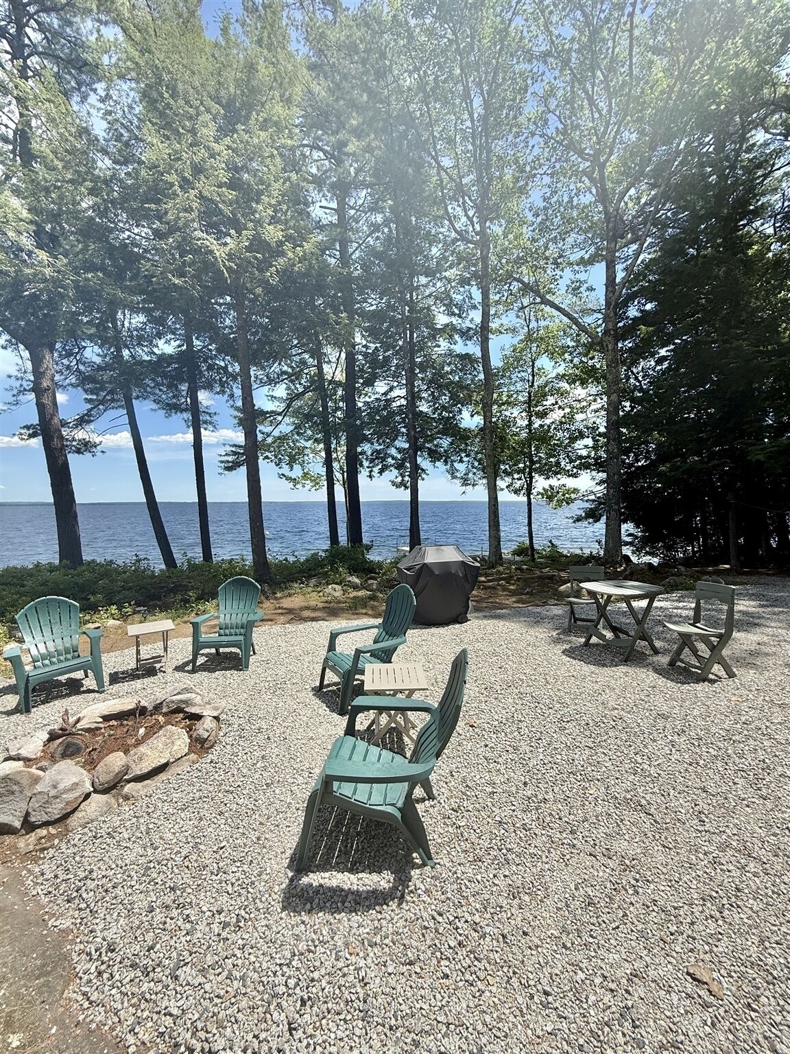 Un espace extérieur aménagé avec des chaises Adirondack, un foyer et une table équipée d’un barbecue, le tout entouré d’arbres et offrant une vue sur une étendue d’eau.