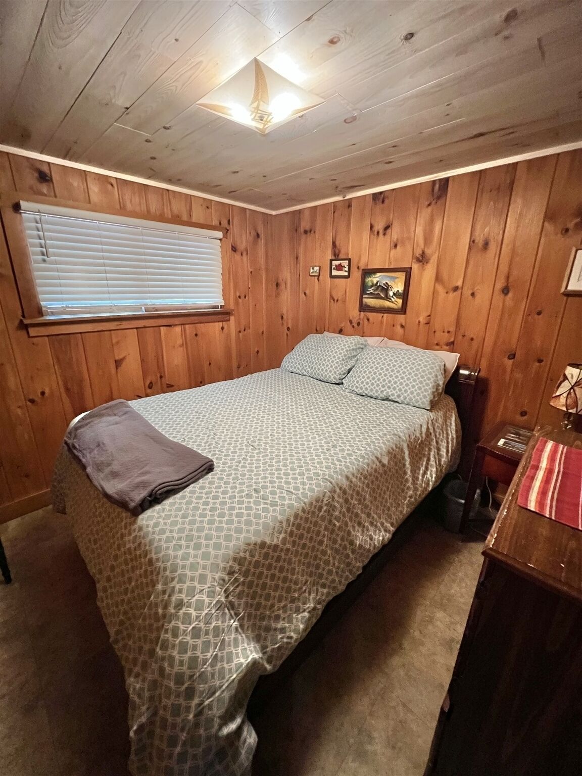 A wooden-paneled room with a bed, a window with blinds, a small table with a lamp, and a picture on the wall.