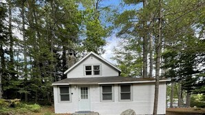 Une petite maison à un étage, dotée d’un toit à pignon, entourée d’arbres et bordée d’une pelouse.