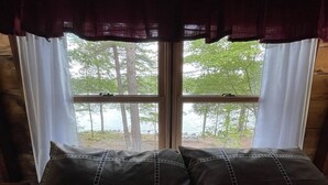 Un lit avec une tête de lit marron foncé et des oreillers assortis, une fenêtre avec des rideaux blancs, et une vue sur les arbres et l’eau à l’extérieur.