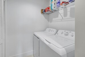 Une buanderie équipée de deux machines à laver à chargement frontal, de couleur blanche, et d’une étagère située au-dessus, où sont rangés divers produits de lessive.
