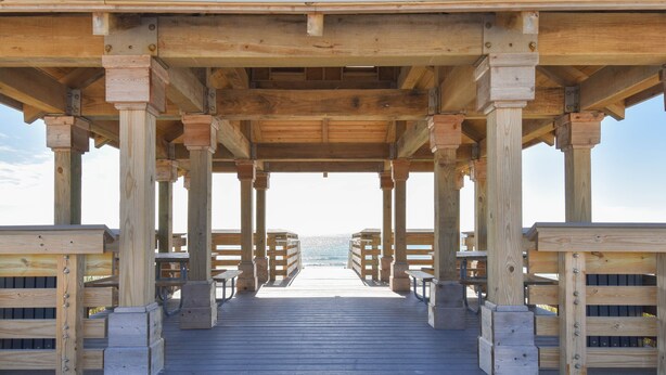 Een houten steiger met uitzicht op de oceaan, ondersteund door kolommen en voorzien van bankjes.