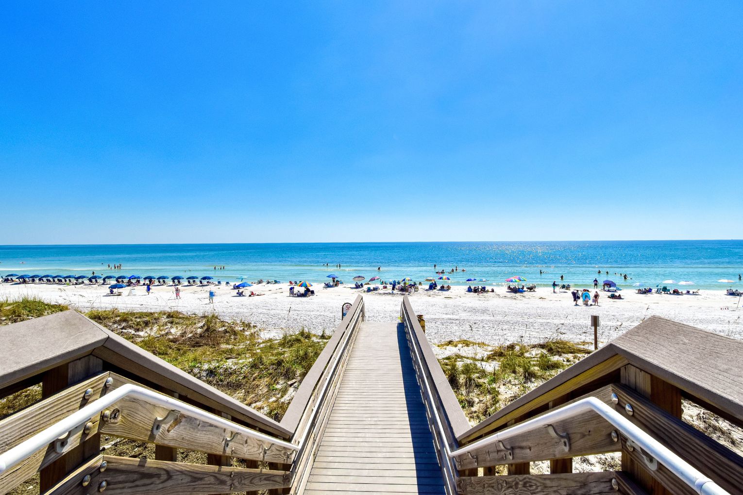 Een houten looppad dat naar een strand leidt, met parasols en mensen.