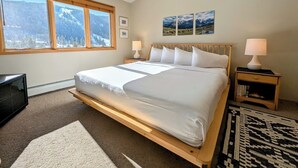 Een slaapkamer met een groot bed, een houten hoofdbord, twee nachtkastjes met lampen, een televisie en een raam met uitzicht op de bergen.