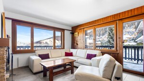 A spacious living area with a sofa set, a wooden coffee table, and large windows offering a view of a snowy mountain landscape. - Arnica 1 - 6 pers - piscine (Haute-Nendaz)