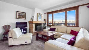 A living room with a fireplace, sofa, TV, and large windows with a mountain view. - Arnica 1 - 6 pers - piscine (Haute-Nendaz)