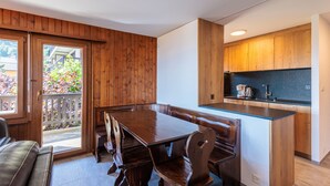 A dining area with a wooden table and chairs, a kitchen with wooden cabinets, and a view of a garden through a glass door. - Arnica 1 - 6 pers - piscine (Haute-Nendaz)