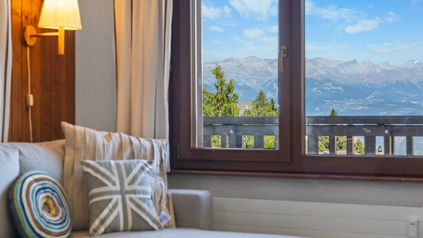 A room with a view of mountains through a window, a sofa with cushions, and a wall-mounted lamp.