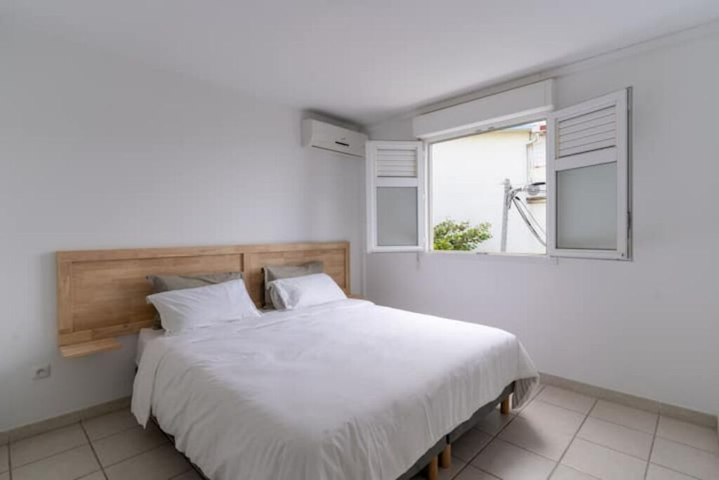 Ein Schlafzimmer mit Holz-Kopfteil, einem Bett mit weißer Bettwäsche, einem Fenster mit Rollläden und einer kleinen Klimaanlage an der Wand.