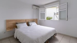 A bedroom with a wooden headboard, a bed with white bedding, a window with shutters, and a small air conditioning unit on the wall. - Kaz Mitan Appt. les Pieds Dans Leau ! (Les Trois-Îlets)