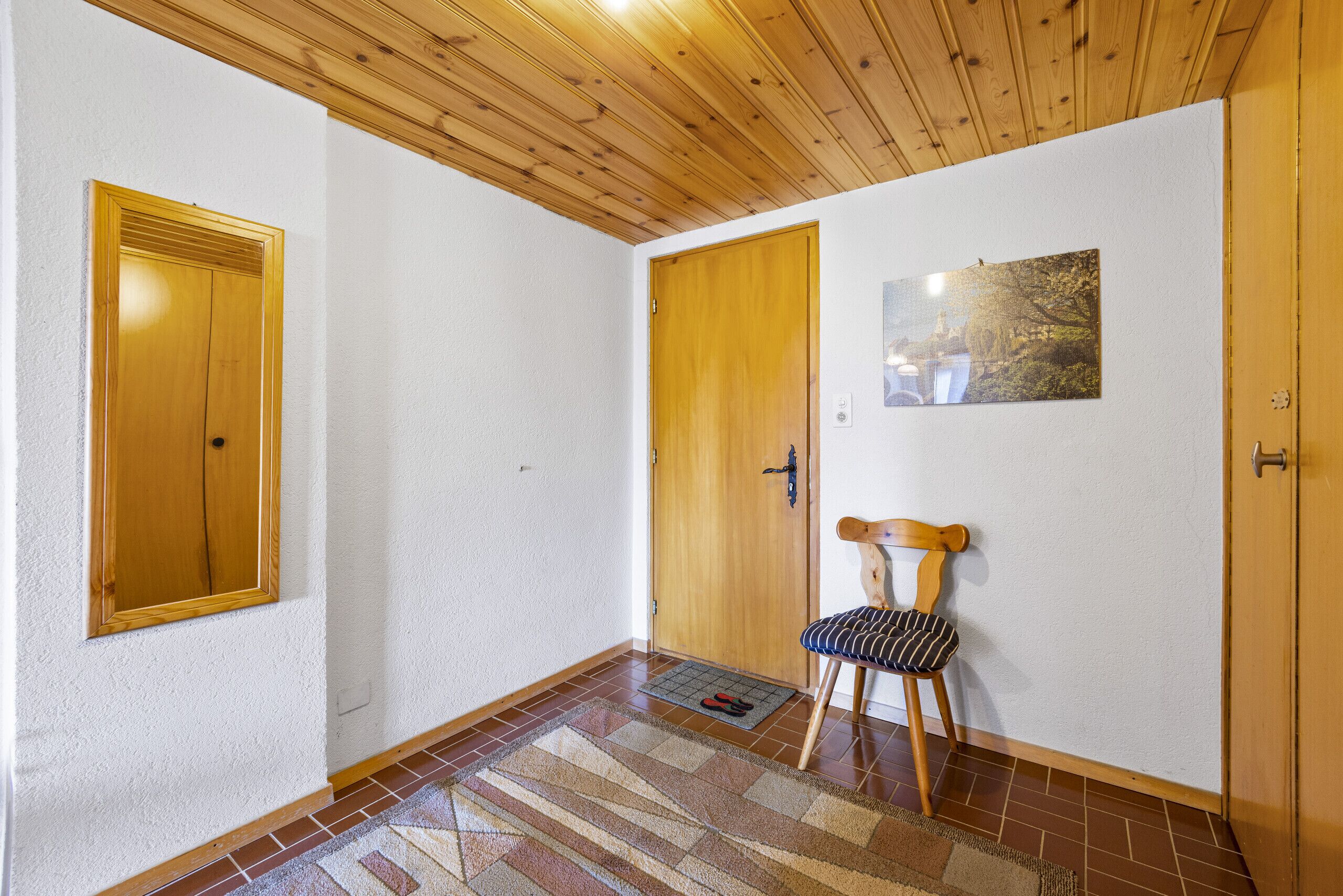 A hallway with wooden flooring, a wooden door, a chair, and a rug.