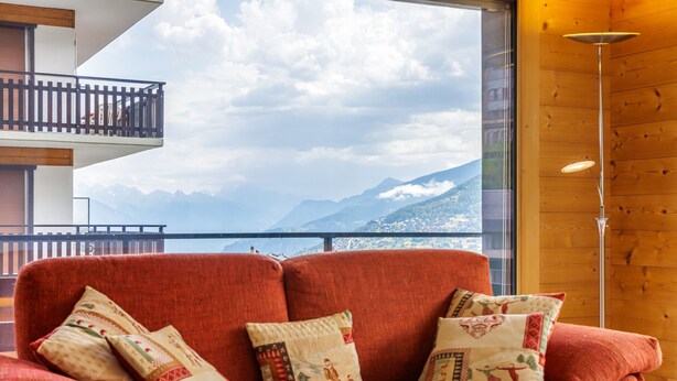 Un canapé rouge avec des coussins à motifs, dans une pièce au décor intérieur en bois, offrant une vue sur les montagnes par une grande fenêtre.