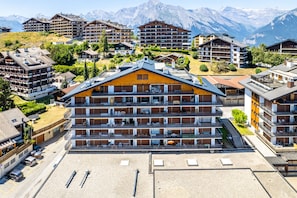 A multi-story hotel with balconies, a parking area, and a mountainous backdrop.