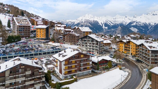 Une ville enneigée, avec des bâtiments à plusieurs étages, un parking et une route qui serpente à travers le paysage.