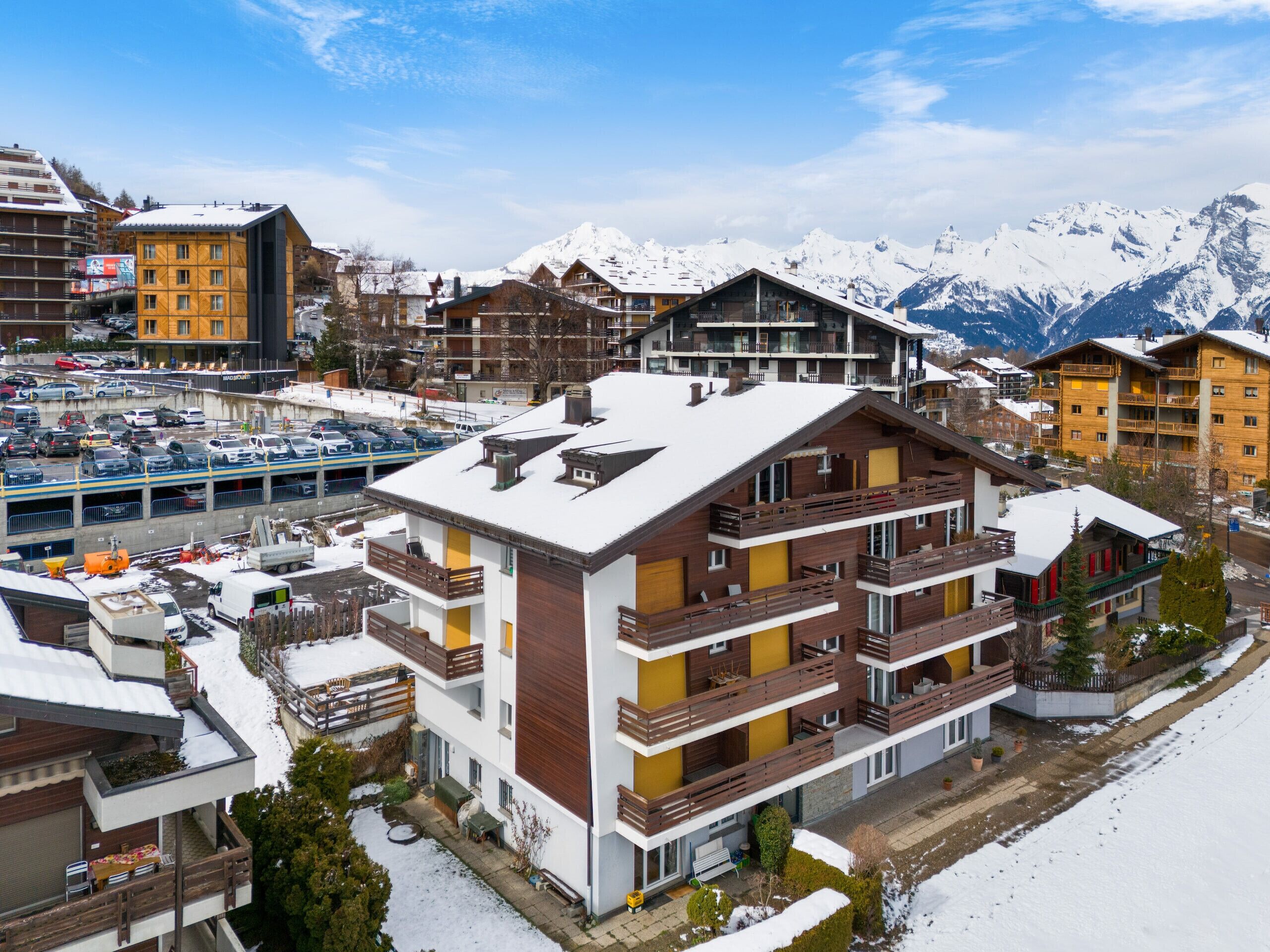 Ein mehrstöckiges Hotel mit Balkonen, einem Parkplatz und verschneiten Bergen im Hintergrund.
