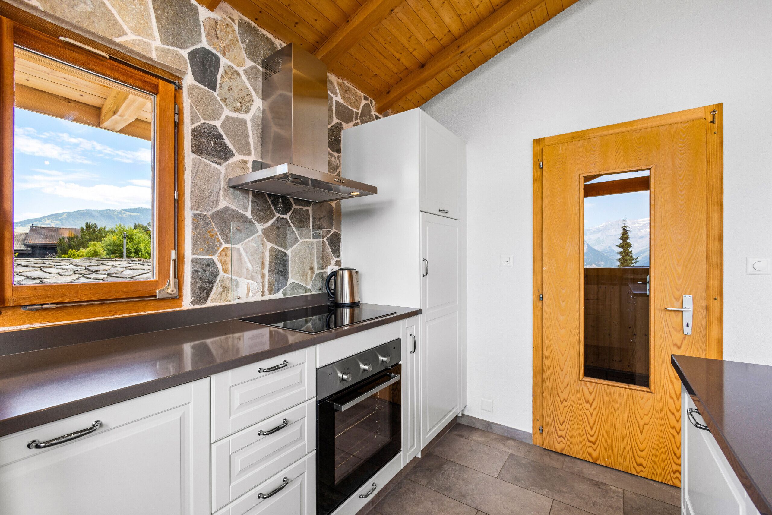 A kitchen with a stone wall, wooden ceiling, and a view of mountains through a window.