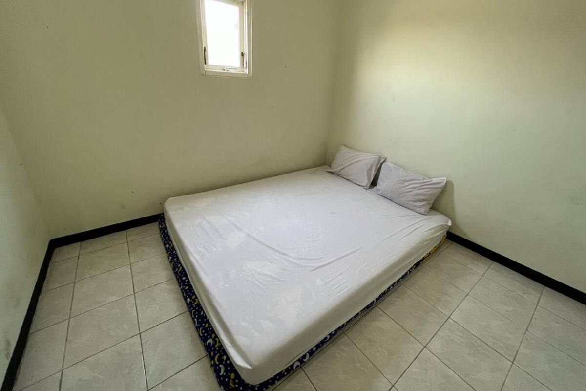 Una habitación pequeña con una cama individual, un colchón blanco sencillo, un ventanal y suelo de baldosas.