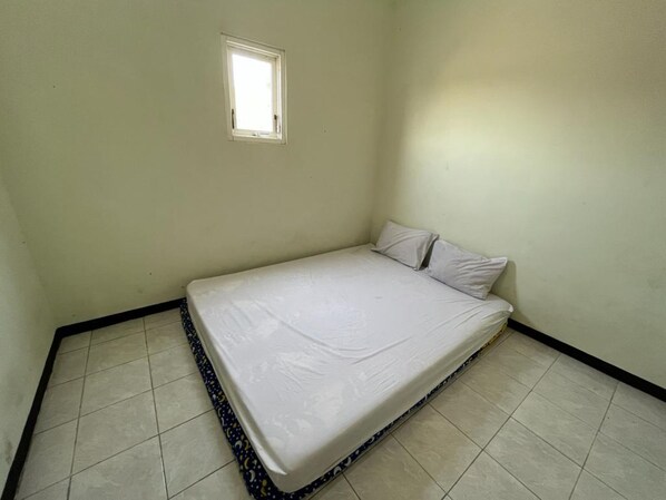 A small, single-bed room with a plain white mattress, a small window, and tiled flooring. - OYO Life  92982 Kost Berkah Ibu (Singasari)