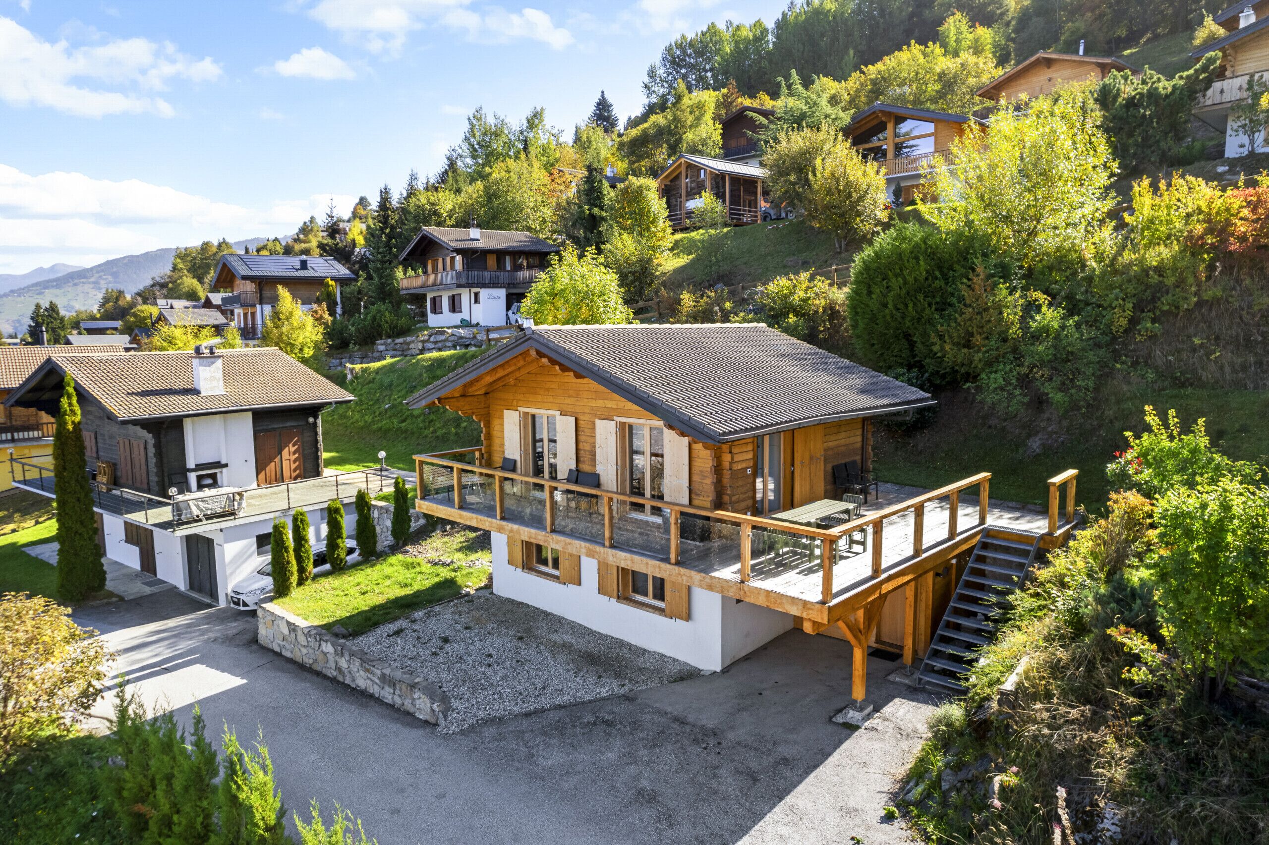Un moderno chalet con terraza de madera y base de piedra, rodeado de exuberante vegetación y otros chalets en una zona montañosa.