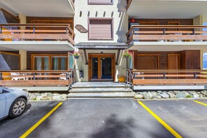 A multi-story building with a wooden balcony, a stone facade, and a parking area.