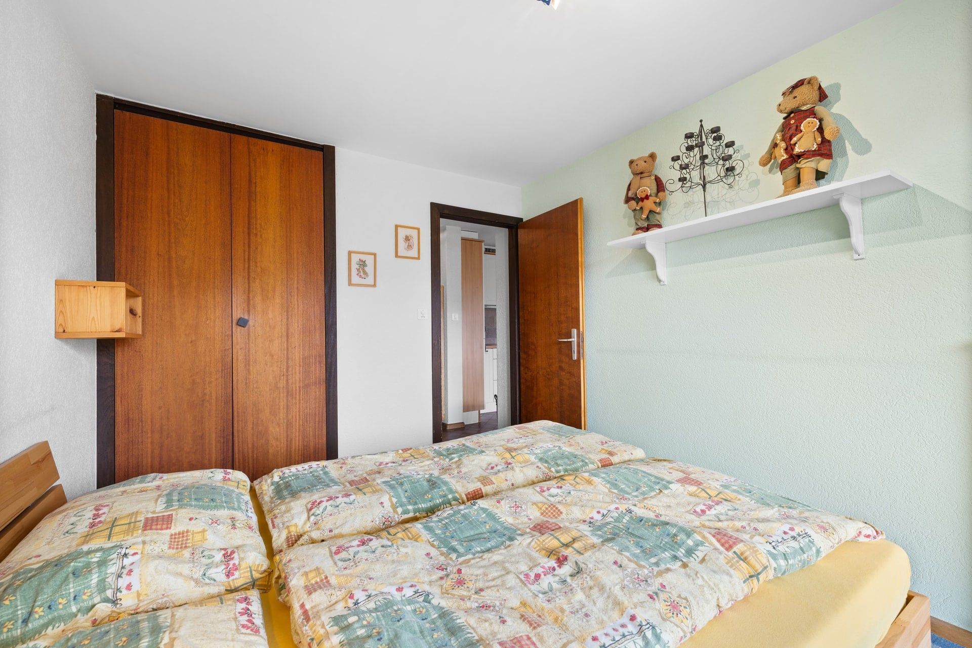 Une chambre avec une armoire en bois, un lit recouvert d’une couette à motifs floraux, une étagère blanche ornée d’ours en peluche et une porte donnant sur une autre pièce.