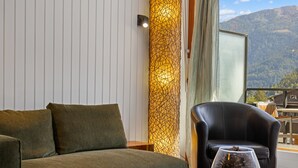 A room with a sofa, a chair, a table with a glass containing pine cones, a patterned wall, and a view of mountains through a window. - Les Platanes E4 - 5 pers - centre station (Haute-Nendaz)