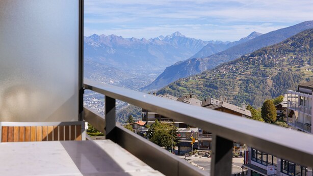 A mountain view from a balcony with a metal railing, overlooking a valley with buildings and mountains. - Les Platanes E4 - 5 pers - centre station (Haute-Nendaz)