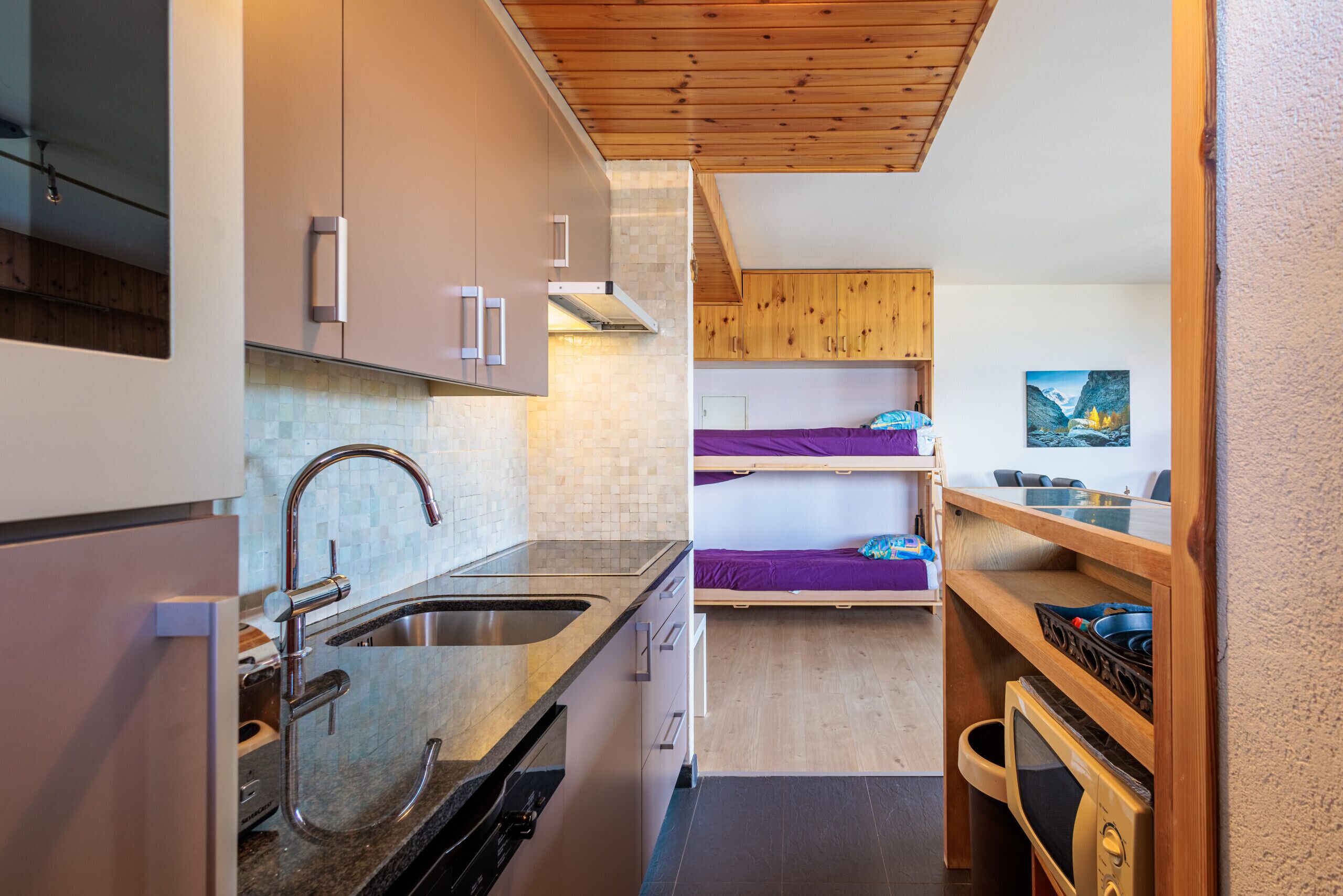 A compact kitchen with a sink, cabinets, and a microwave. There is a bunk bed with a painting on the wall.