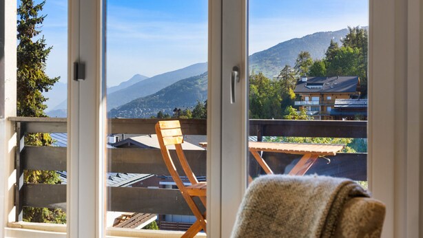 A room with a large window, wooden deck, and a view of mountains.