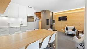 Une cuisine moderne avec des armoires blanches, une table à manger en bois et des chaises blanches. On y trouve un four encastré et un mur en bois sur lequel est fixée une télévision.