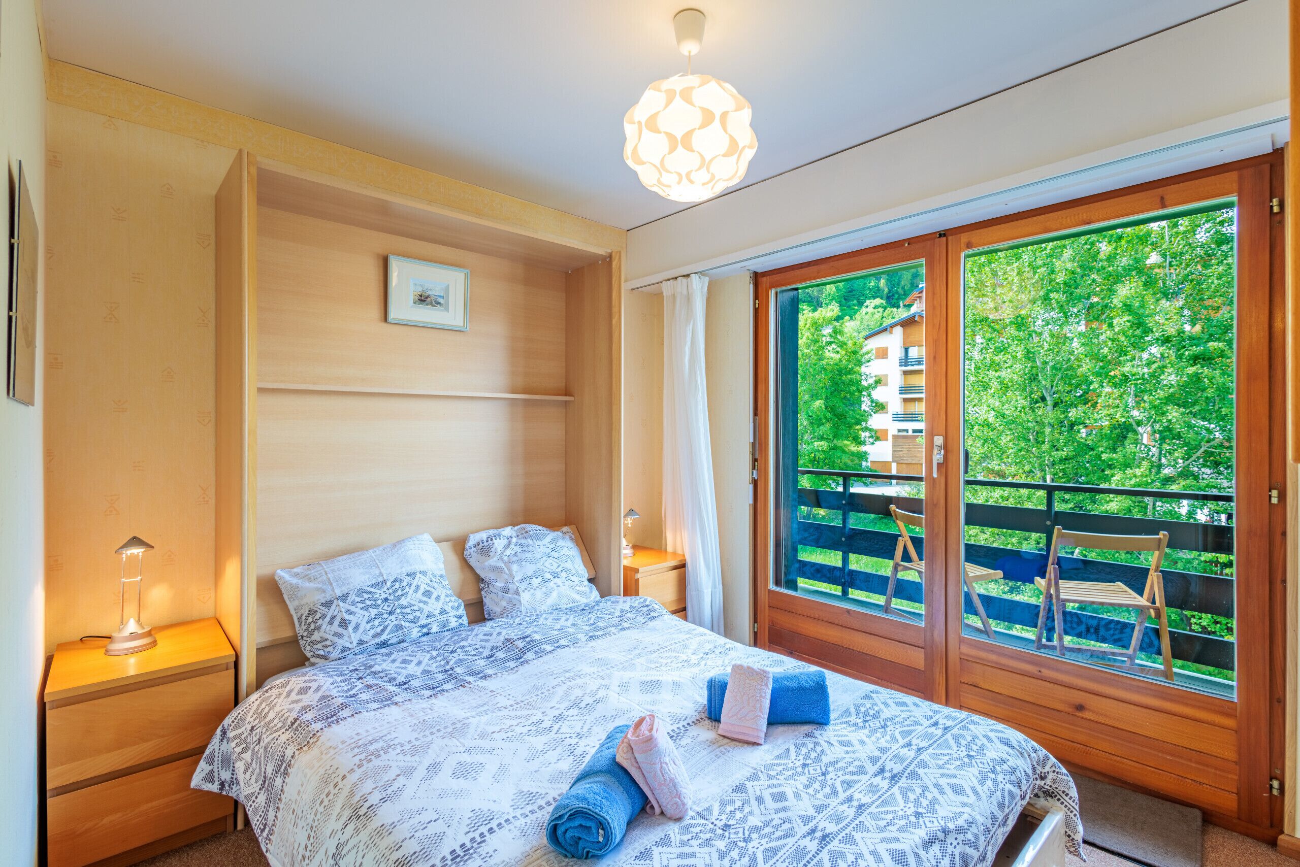 A bedroom with a bed, bedside tables, a lamp, a window with a view of greenery, and a ceiling light.