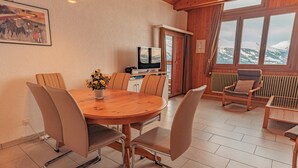 A dining area with a wooden table and chairs, a television, and a view of snow-capped mountains through large windows.