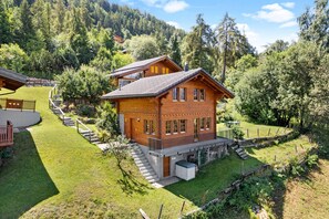 A wooden cabin with a balcony, surrounded by a well-maintained lawn and a stone wall.