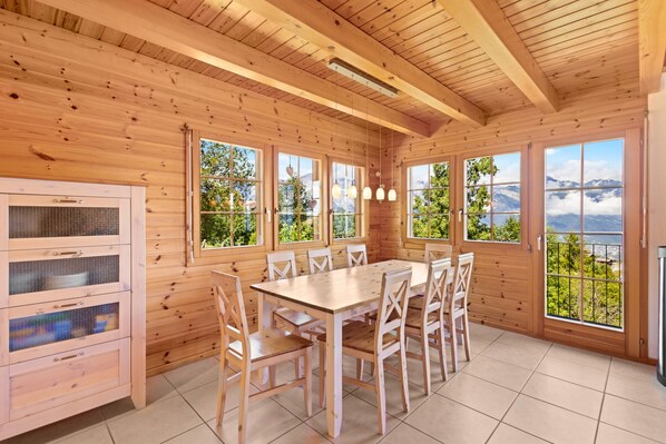 A wooden cabin with a dining table and chairs, large windows with a mountain view, and a kitchen area with cabinets and a refrigerator.