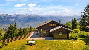 A wooden cabin with a deck, surrounded by lush greenery and mountains in the background. - Gisèle - 8 pers - près des pistes (Haute-Nendaz)