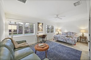 A spacious living room with a leather couch, a round ottoman, a bed with patterned bedding, and a desk with a chair. - Pet-Friendly Inland Home | Resort Style Yard & Pool (Marco Island)