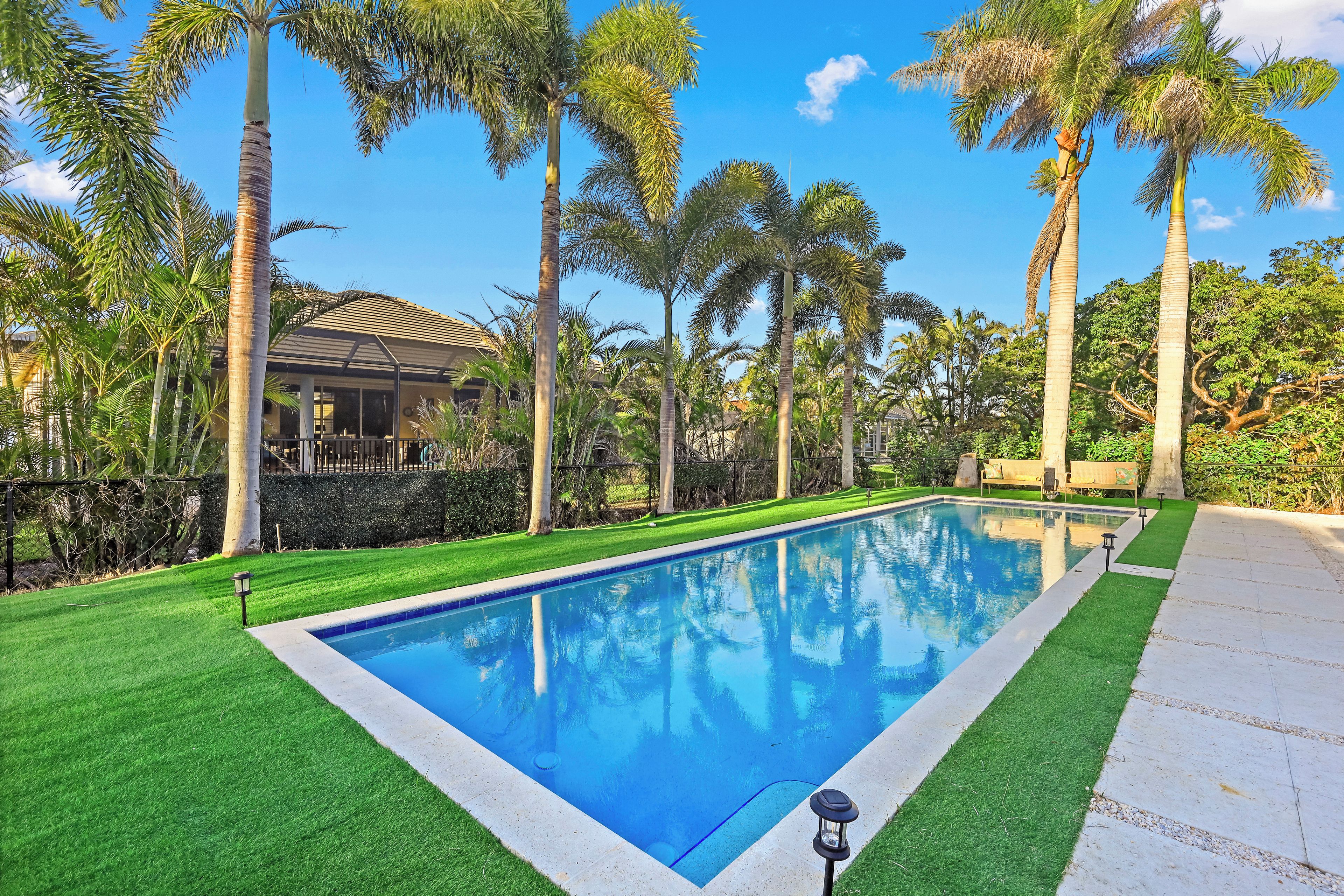 A rectangular swimming pool with a clear blue water surface, surrounded by well-maintained artificial grass, palm trees, and a house in the background.