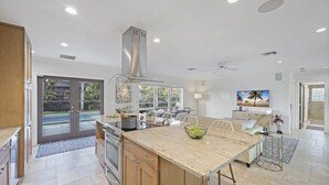 A modern kitchen with a large island, a dining area with a TV, and a view of the outdoors. - Pet-Friendly Inland Home | Resort Style Yard & Pool (Marco Island)