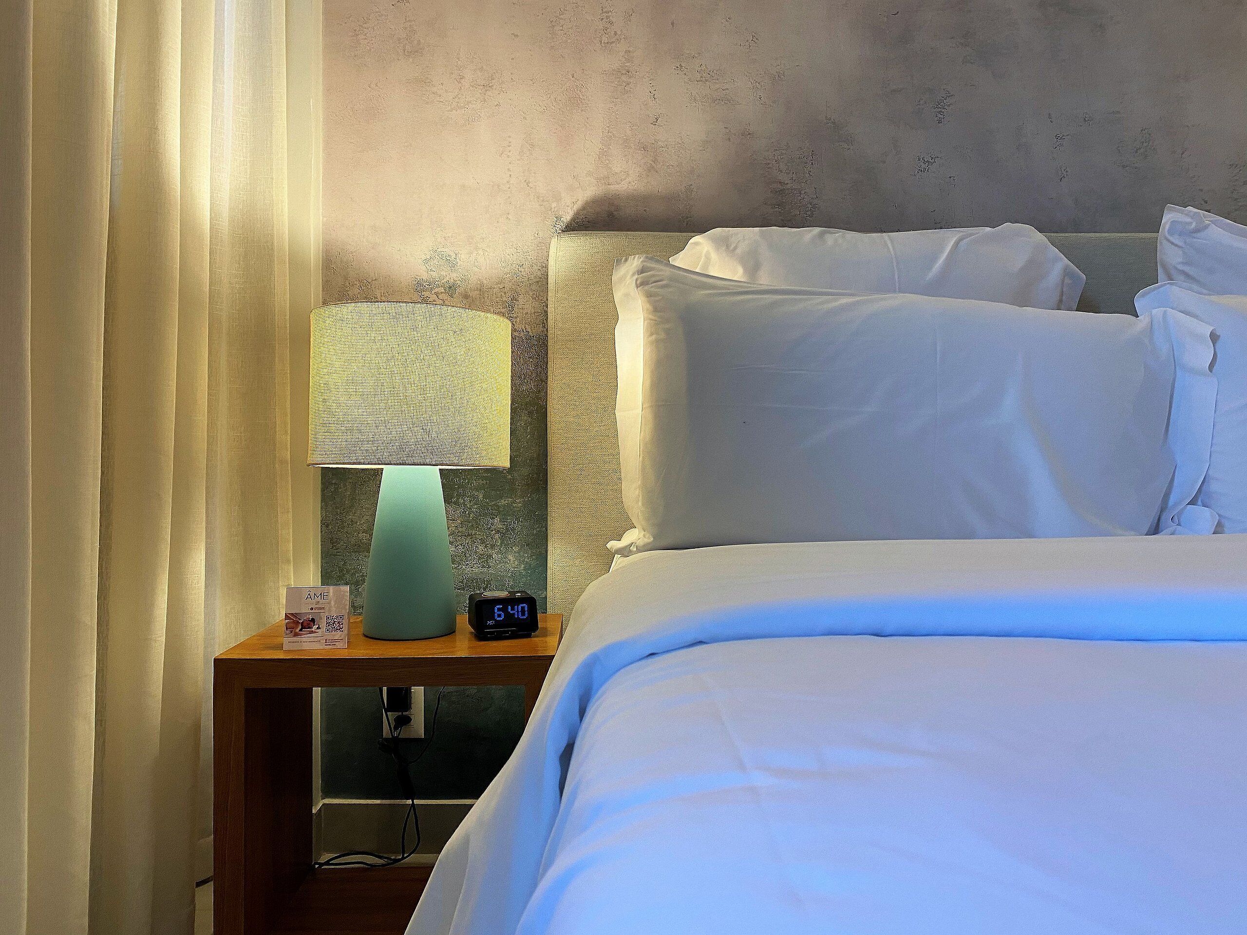 A hotel room with a bed, white linens, a nightstand with a lamp, and a clock displaying the time.