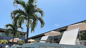 Een zwembadgedeelte met ligbedden en parasols, omgeven door palmbomen en moderne gebouwen.