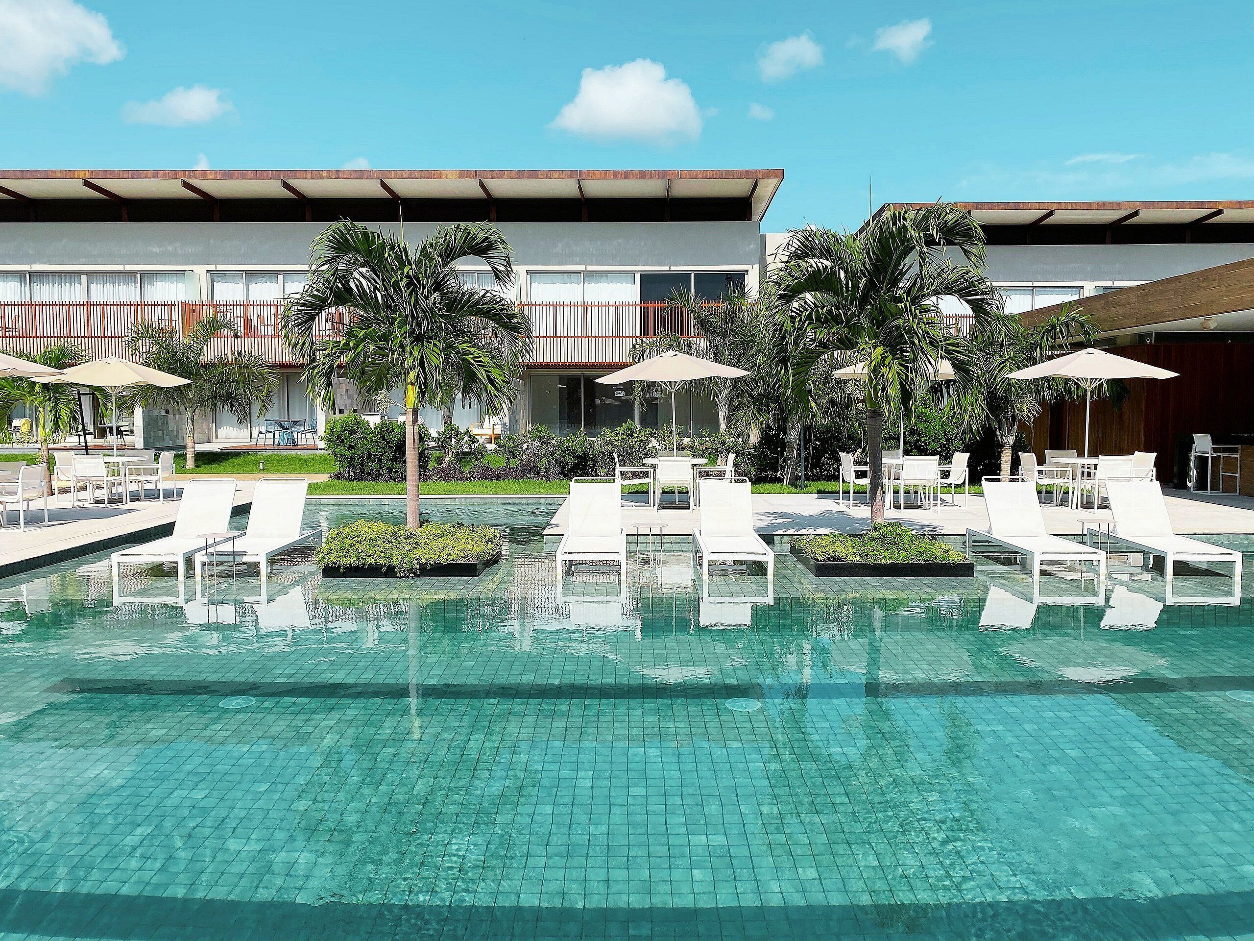 A pool area with lounge chairs and umbrellas, surrounded by palm trees and a building with a modern design.