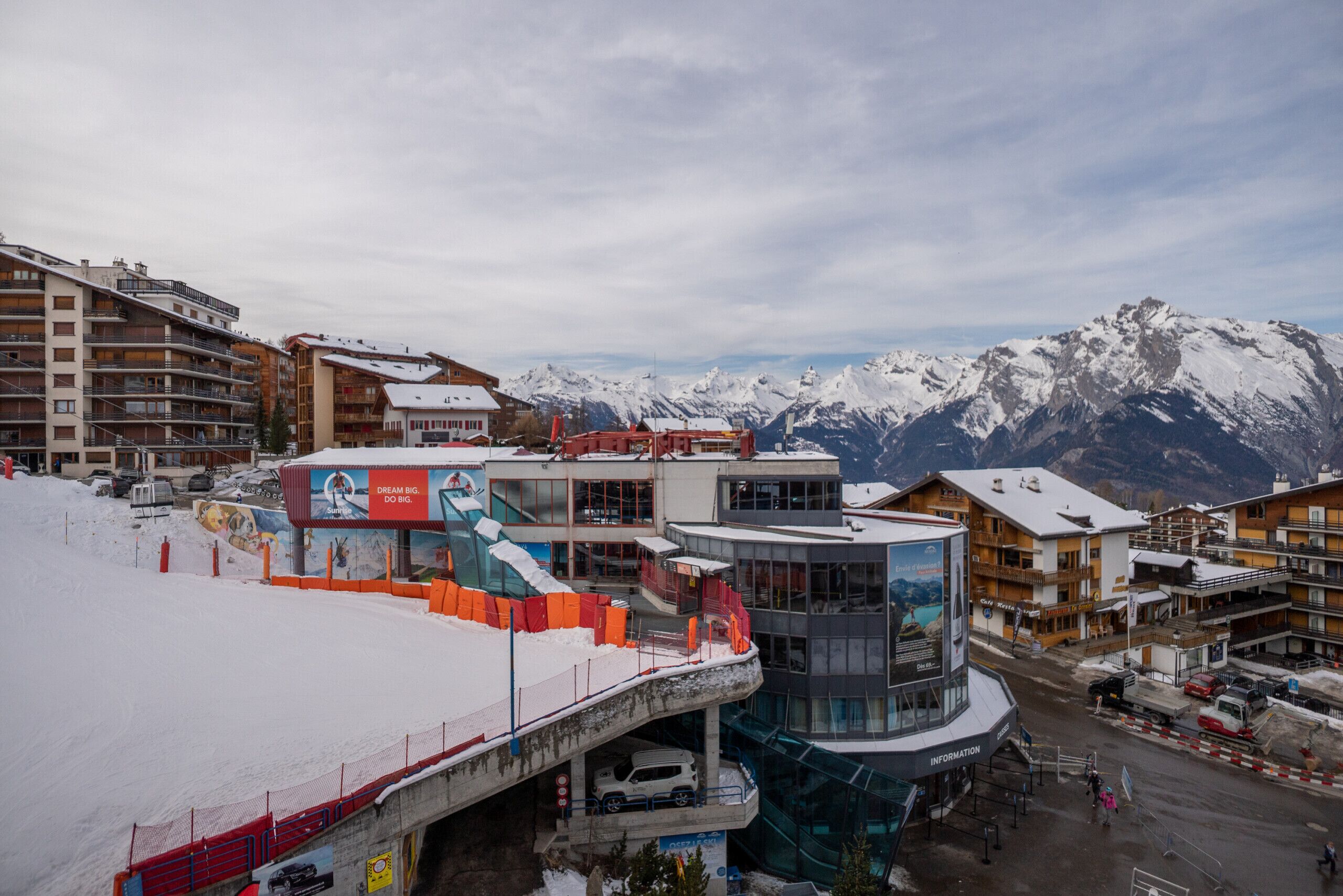 A ski resort with buildings, a covered area, and snow-covered mountains in the background.